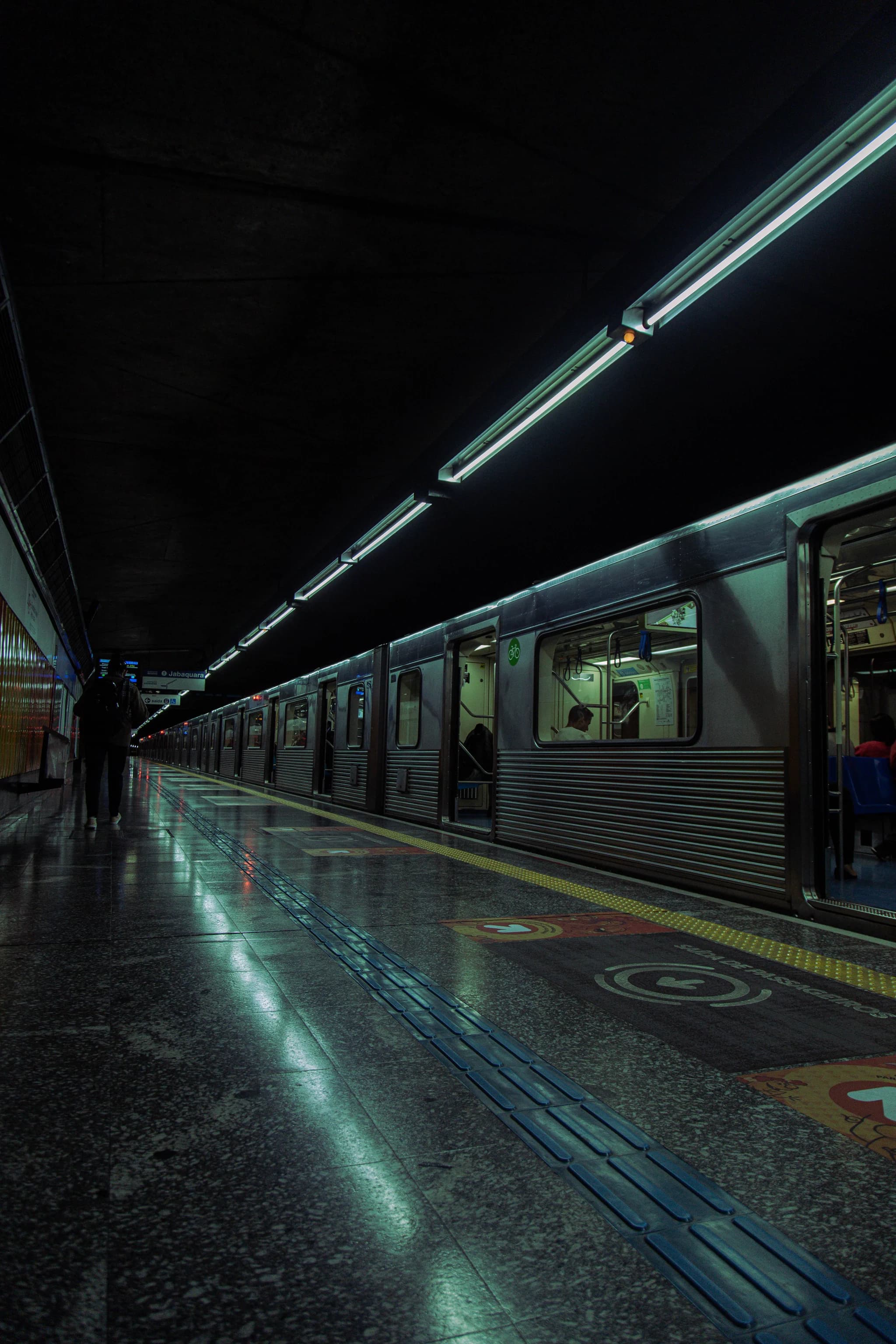 A lone figure waits at the end of an empty subway platform, sodium light on wet tile, long lens compression, heavy film grain